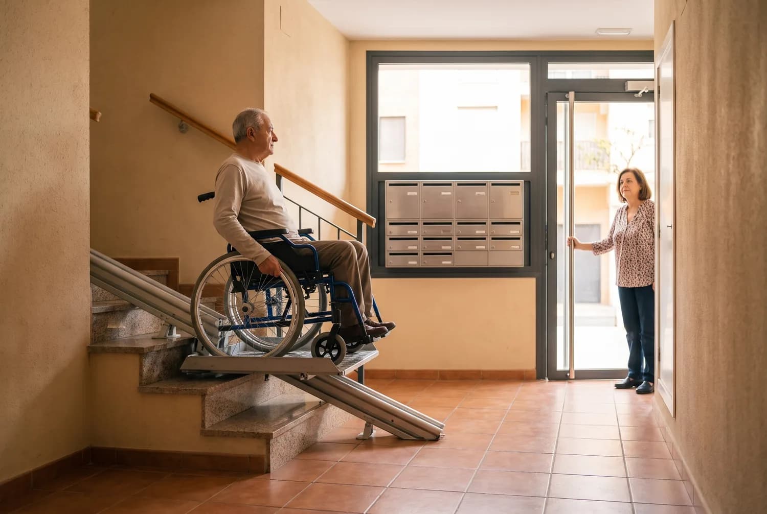 Un hombre en silla de ruedas sube sobre una plataforma salvaescaleras por los peldaños del portal de un edificio mientras una vecina sujeta la puerta (imagen generada con IA)