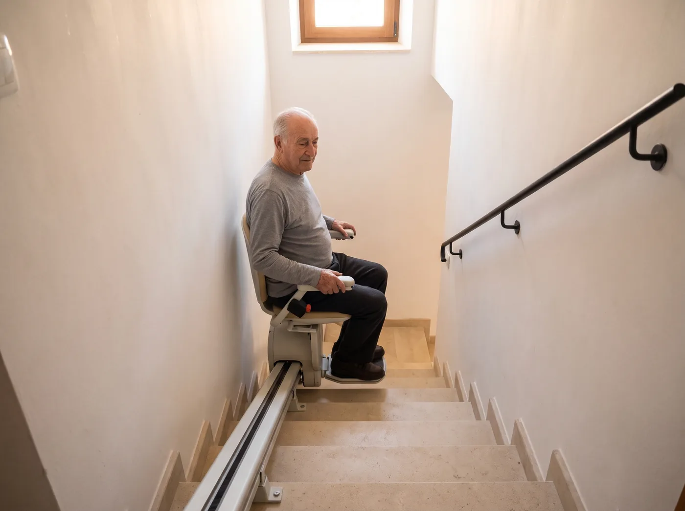 Un hombre mayor baja sentado en una silla salvaescaleras por una escalera recta y estrecha (imagen generada con IA)